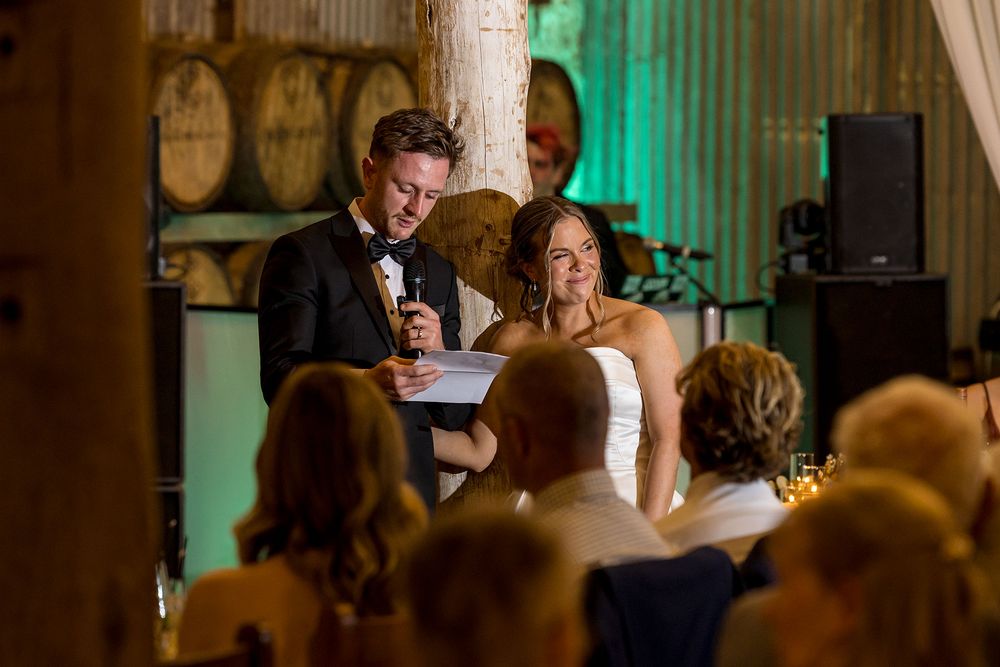 Groom in a tuxedo reads a speech beside his bride in a white gown as wedding guests watch in a rustic barrel room venue.