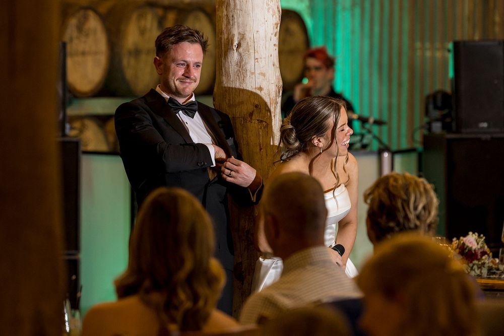Smiling groom in a black tuxedo and bow tie stands beside his bride in white as wedding guests look on in a rustic barn venue.
