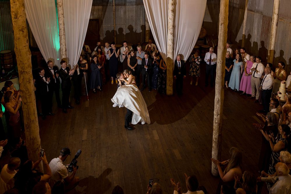 Bride and groom share a first dance on a wooden floor as wedding guests watch from the sides of a rustic venue with draped white fabric.