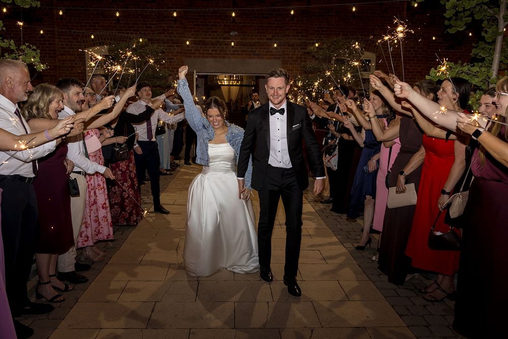 Bride in white gown and groom in tuxedo walk hand in hand between cheering wedding guests holding sparklers at an evening sparkler send-off.