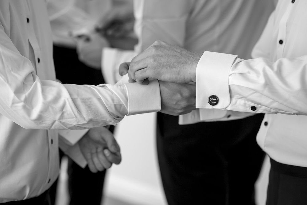 Black and white photo of groomsmen in white dress shirts helping each other fasten cufflinks while getting ready for a wedding.