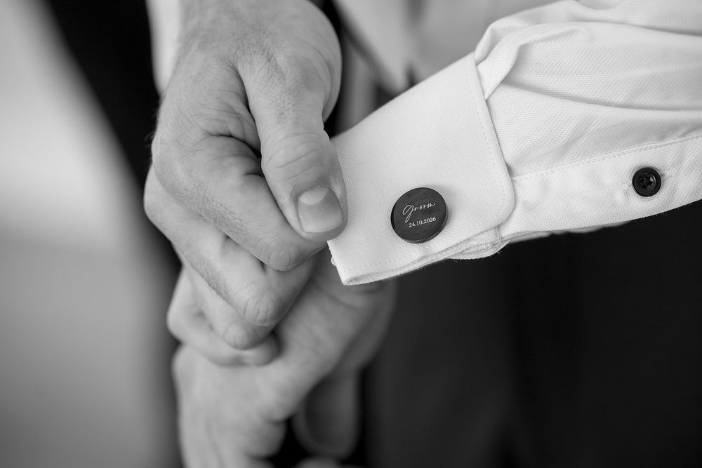Black and white close-up of the Groom's hands fastening a round engraved cufflink on the cuff of a crisp white dress shirt during wedding .