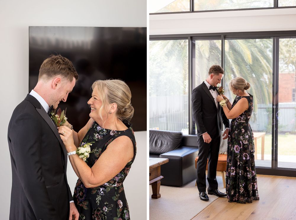 Mother in a floral dress pins a boutonniere on a groom in a black suit, then fixes his lapel flower in a bright room with large windows.
