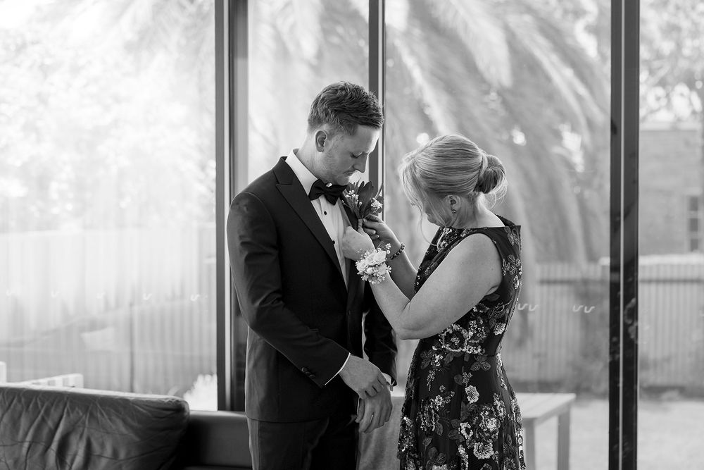 Mother in a floral dress pins a boutonniere on a groom in a black suit, then fixes his lapel flower in a bright room with large windows.