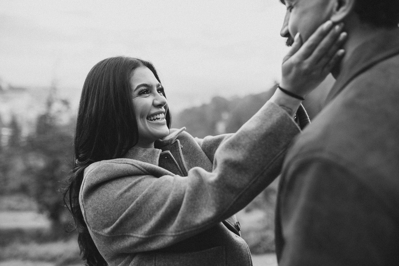Black and white photo of a smiling woman with long dark hair gently touching a man's face in a romantic outdoor couple portrait.