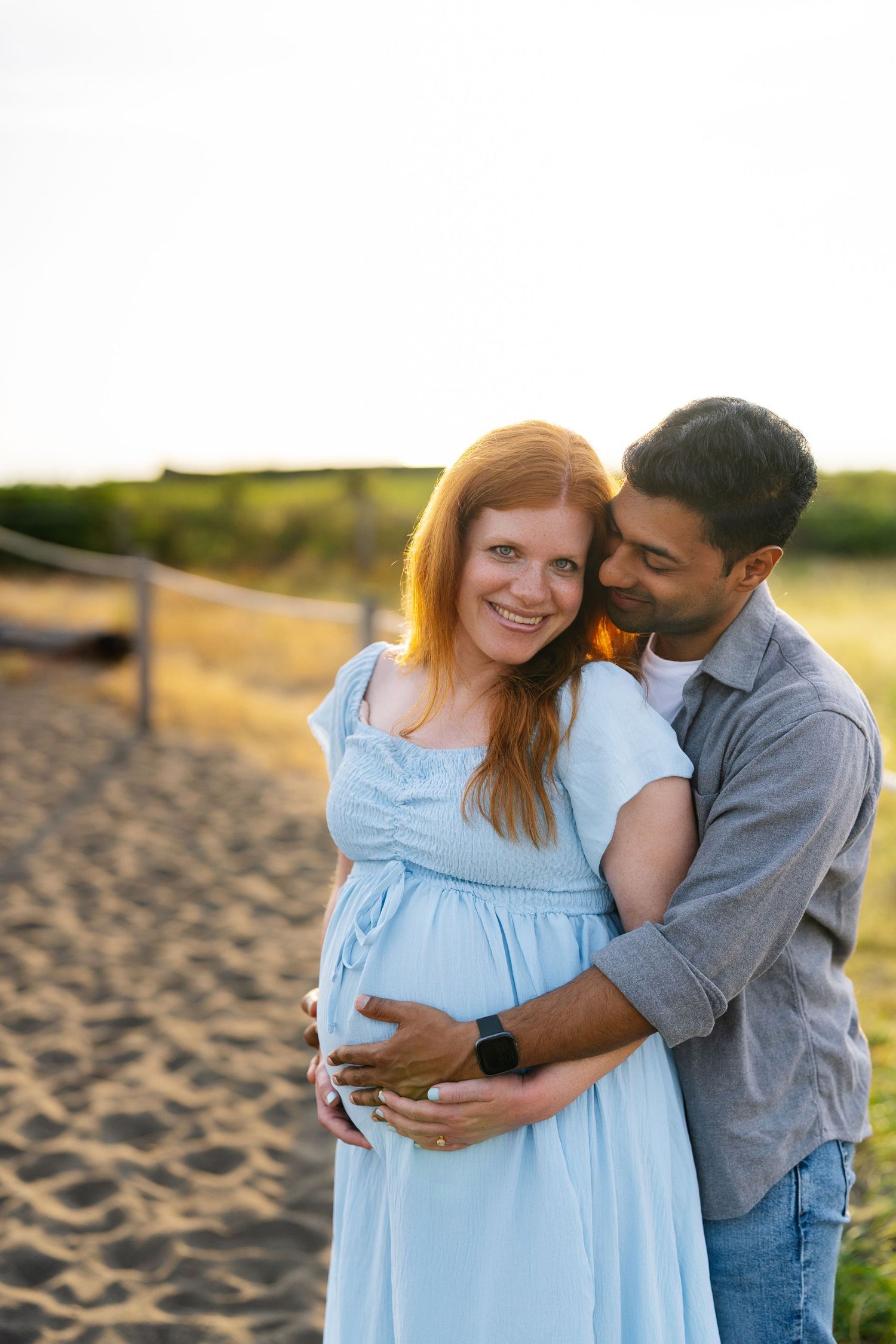 Smiling pregnant woman in a light blue dress embraced from behind by her partner during a golden hour maternity photoshoot on a sandy beach.