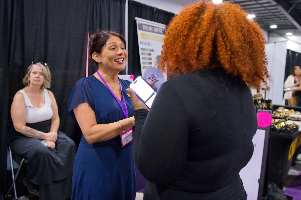 Smiling woman in a blue dress holds a tablet while chatting with a curly-haired woman at a trade show booth.