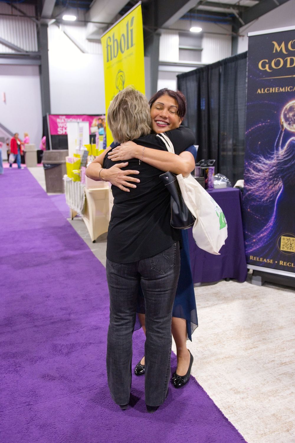 Two women share a warm hug on a purple carpet at an indoor expo, with vendor booths and colorful banners in the background.
