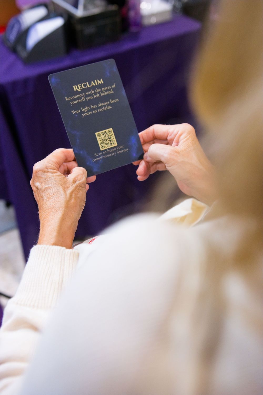 Older woman in a cream sweater holds a dark blue Reclaim card with a QR code at a purple-draped table.