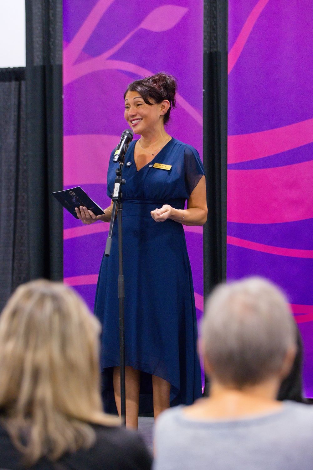 Smiling woman in a navy blue dress speaks into a microphone while holding a tablet before an audience, with a purple banner behind her.