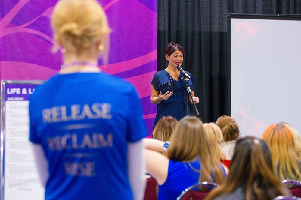Woman speaking into a microphone to a seated audience while a volunteer in a blue Release Reclaim Rise shirt looks on.