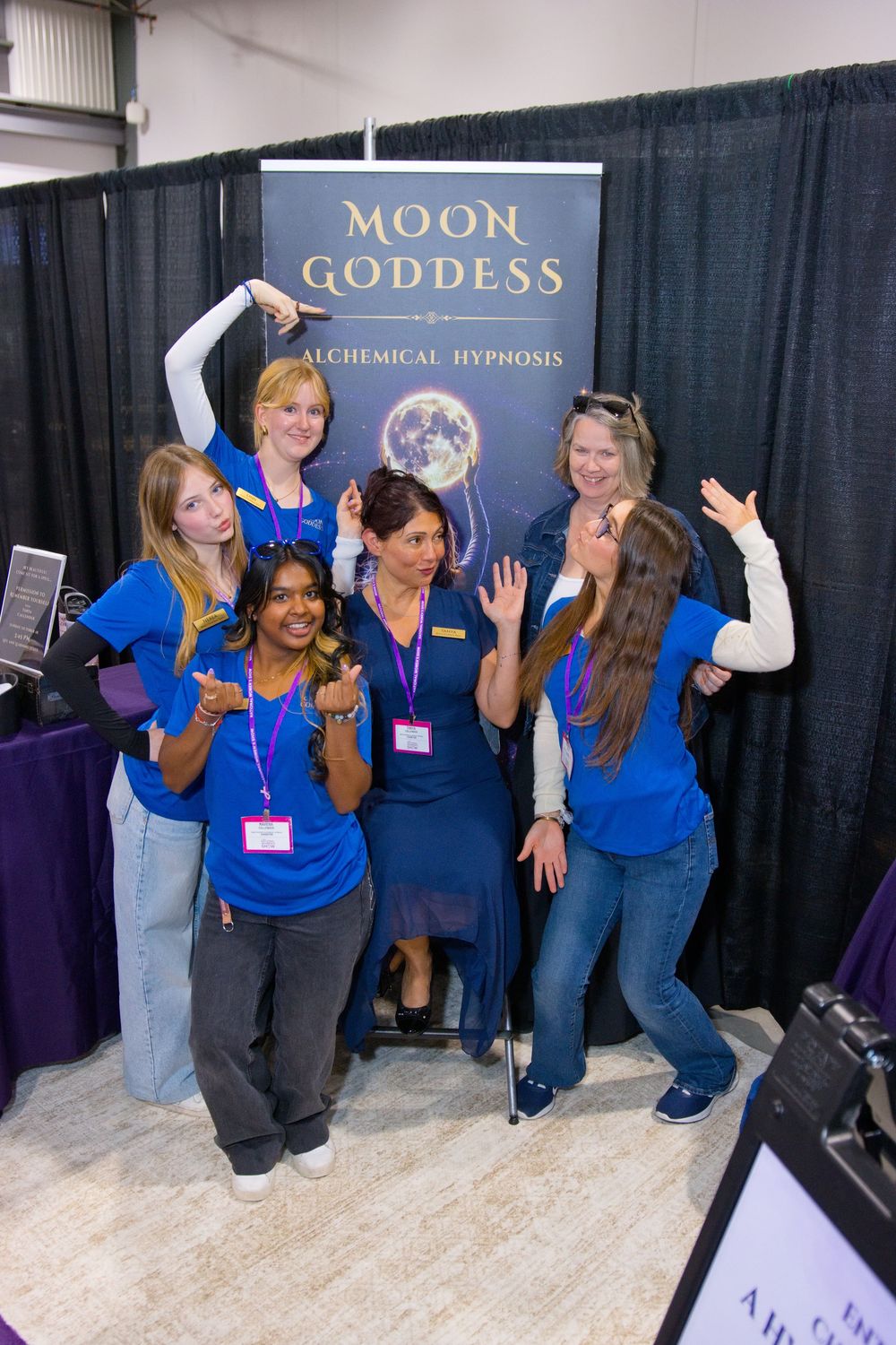 Six women in blue shirts and lanyards pose playfully in front of a Moon Goddess Alchemical Hypnosis banner at an expo booth.