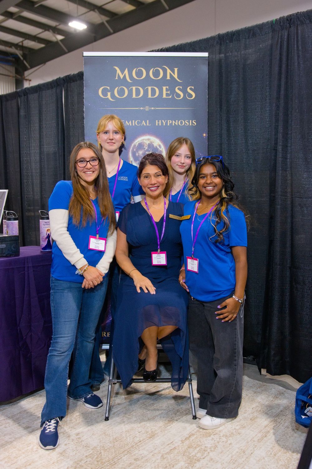 Five women in blue shirts and lanyards pose at a Moon Goddess hypnosis expo booth, one seated in a navy dress.