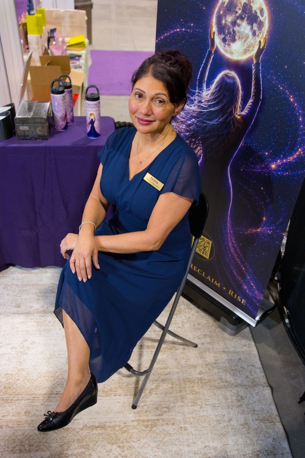Woman in a blue dress and name badge sits on a folding chair at an expo booth beside a purple table and a moon banner.