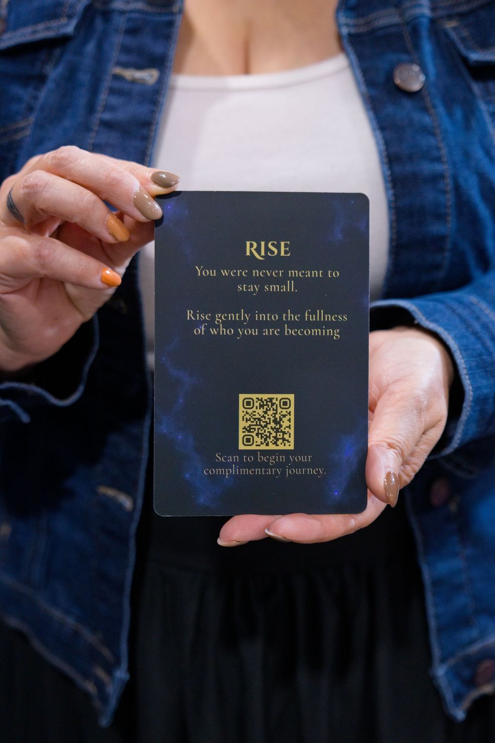 Woman in a denim jacket holds a navy inspirational card reading RISE with a QR code and an encouraging message about personal growth.