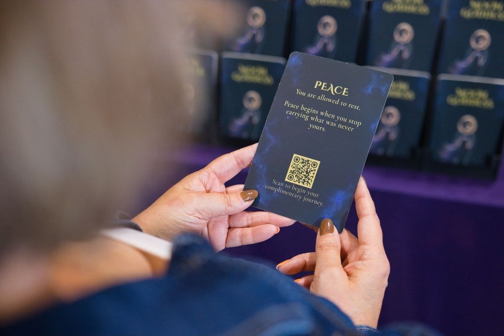 Person holding a navy card reading Peace with a gold QR code, with stacks of matching blue cards displayed on a purple table behind.
