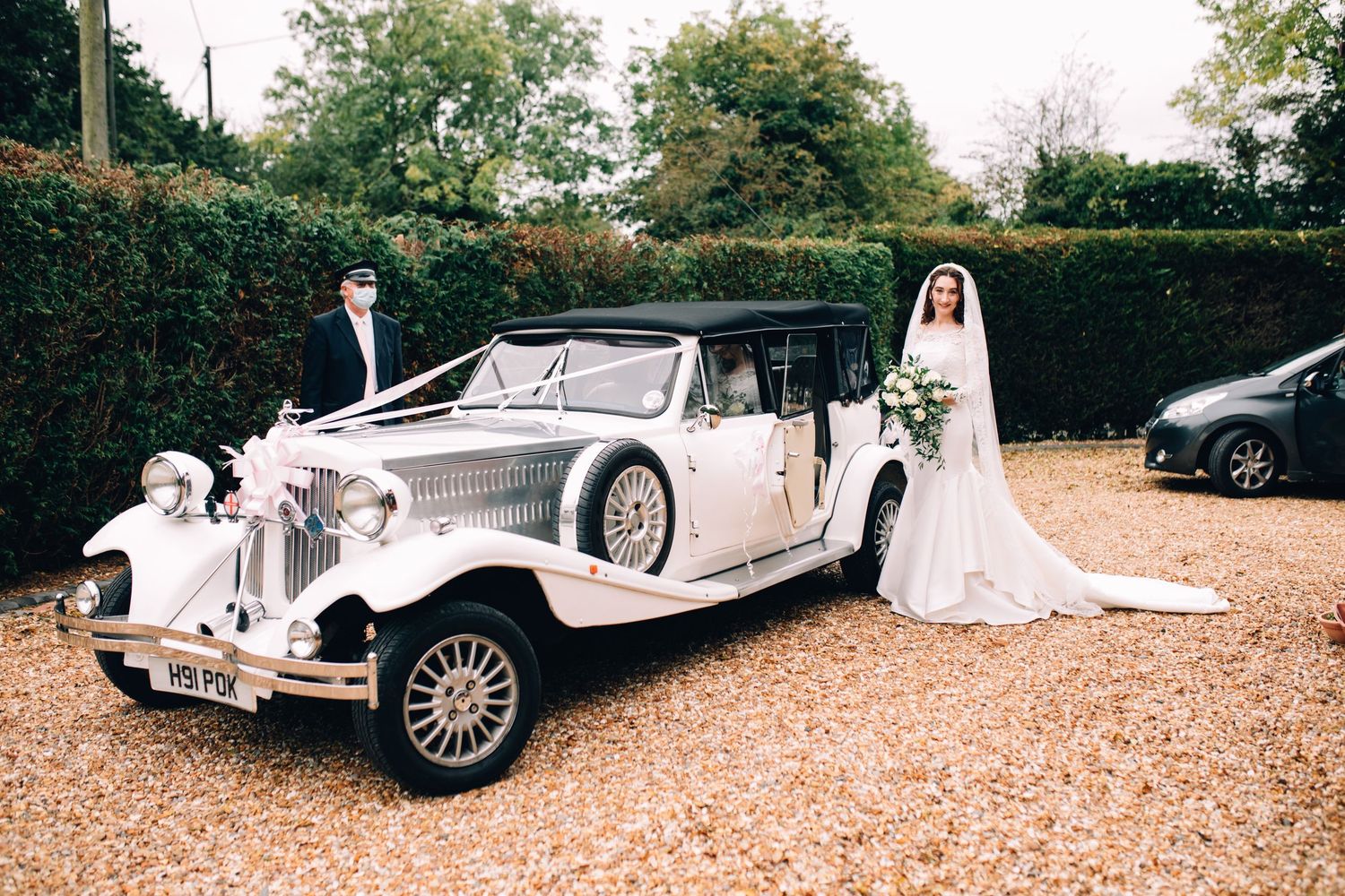 Bride in a white gown holding a bouquet stands beside a vintage cream wedding car while a chauffeur waits on a gravel driveway.
