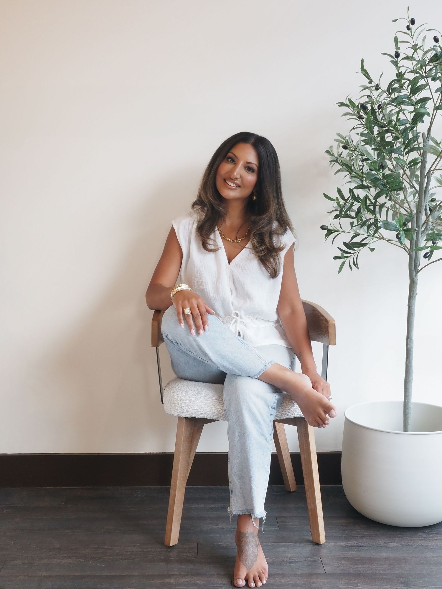 Rhiannon in a casual white attire sits on a couch holding a professional camera while smiling warmly.