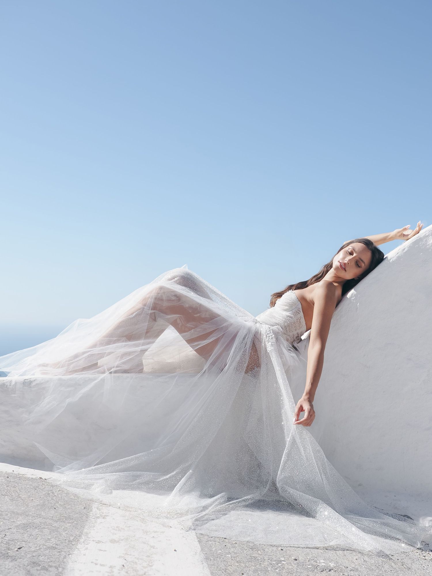Rhiannon in a casual white attire sits on a couch holding a professional camera while smiling warmly.