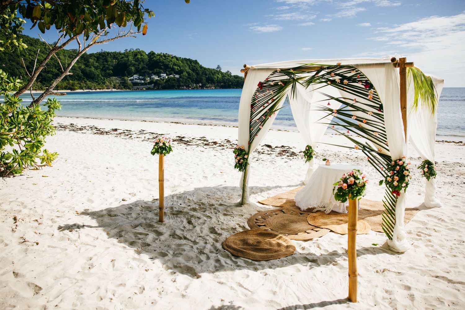 Elegant white canopy wedding arch decorated with tropical greenery and flowers on a sandy beach.