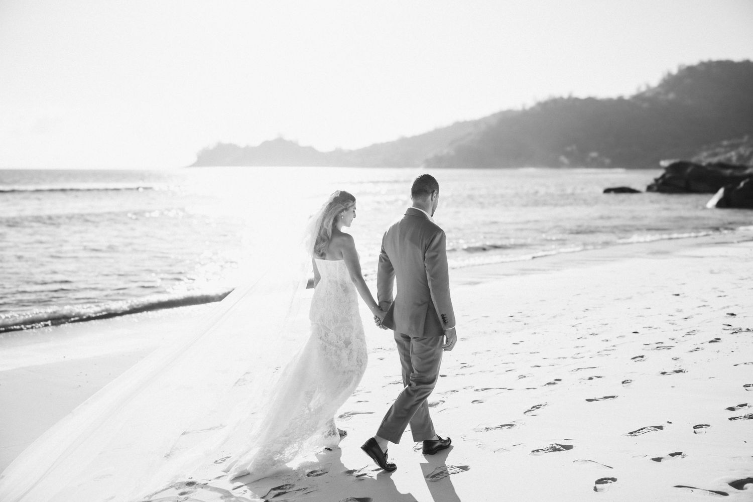 Black and white photo of newlyweds walking hand in hand along sunlit beach shoreline.