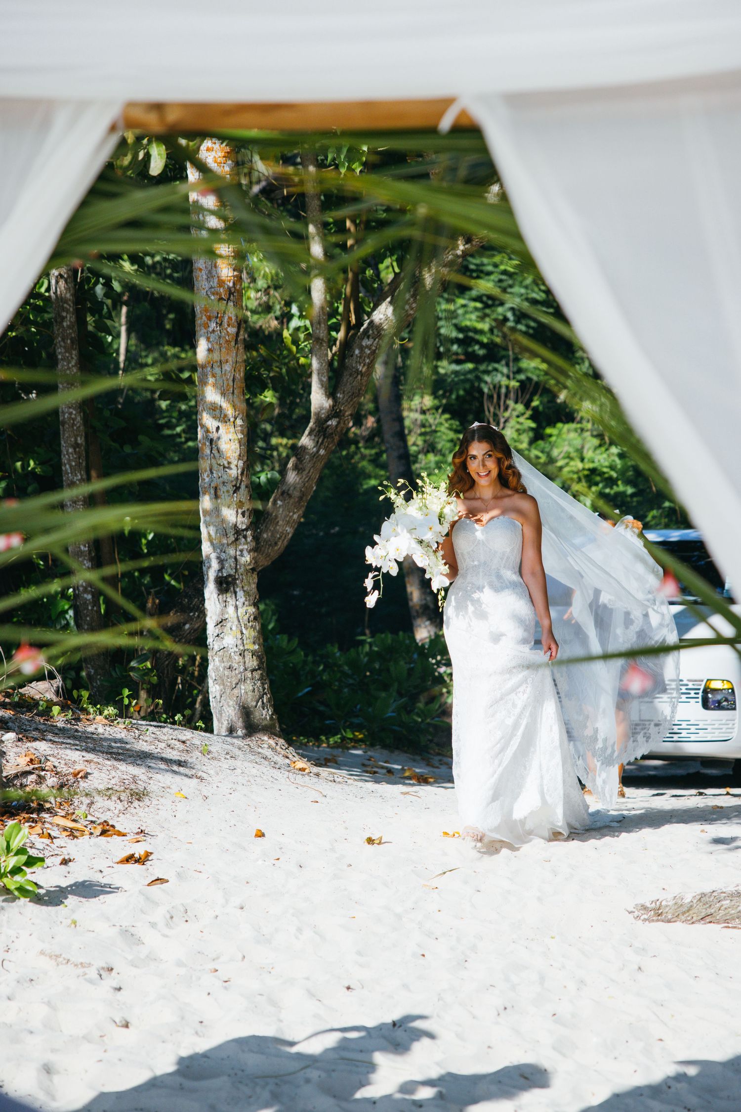 Bride in white strapless gown with veil and orchid bouquet walks toward beach wedding ceremony arch.