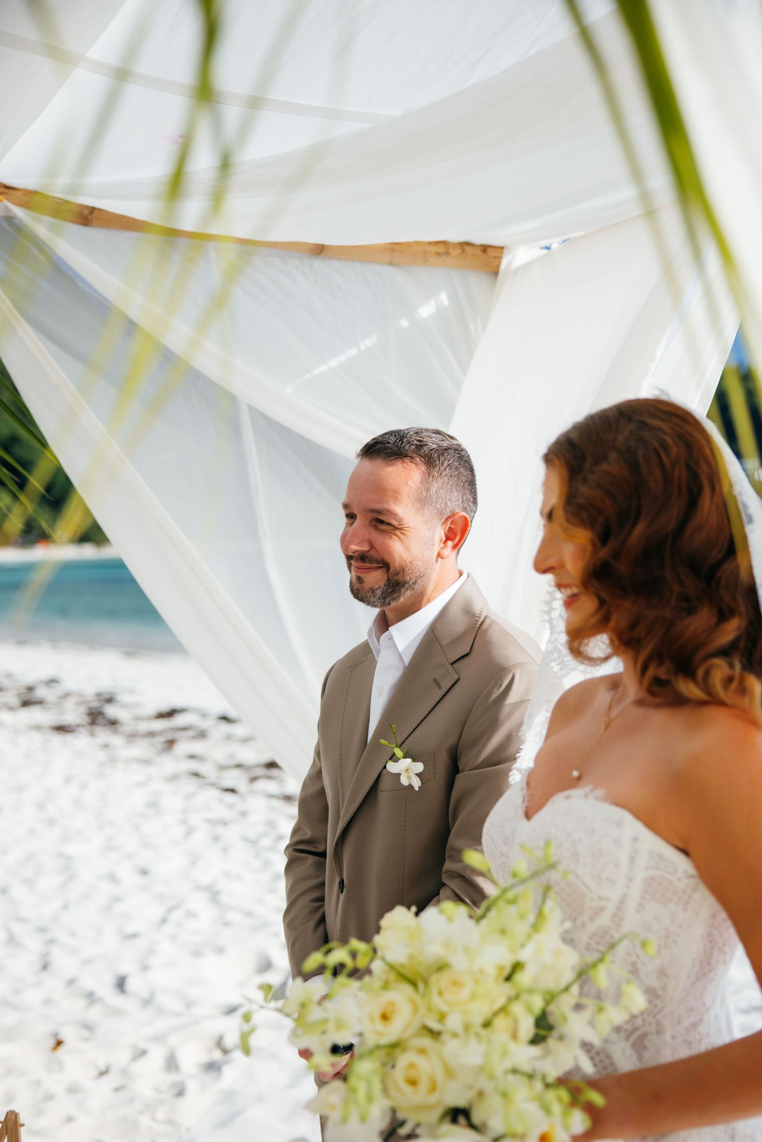 Smiling groom in tan suit stands with bride holding white floral bouquet under a beach wedding canopy.
