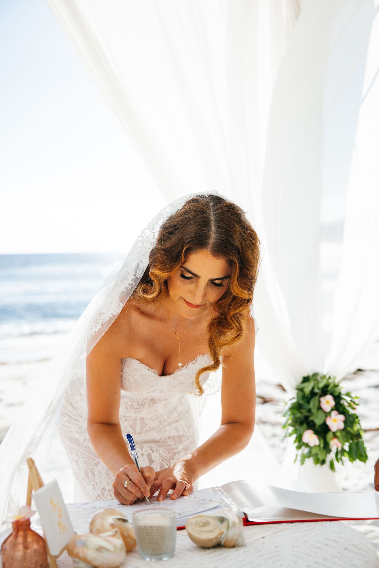Elegant bride in lace wedding dress signing marriage certificate at beach ceremony table.