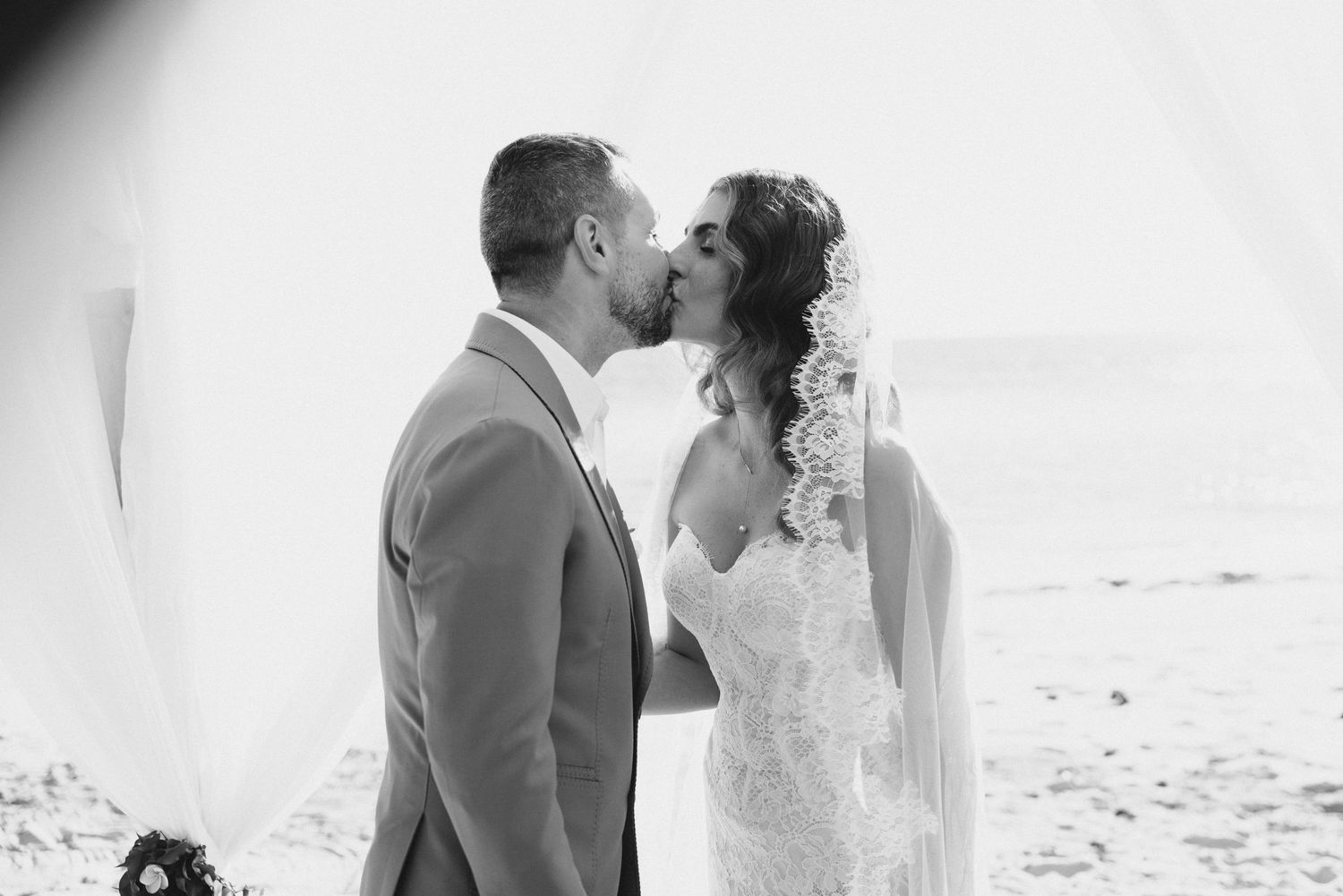 Black and white photo of bride and groom sharing first kiss at beach wedding ceremony.