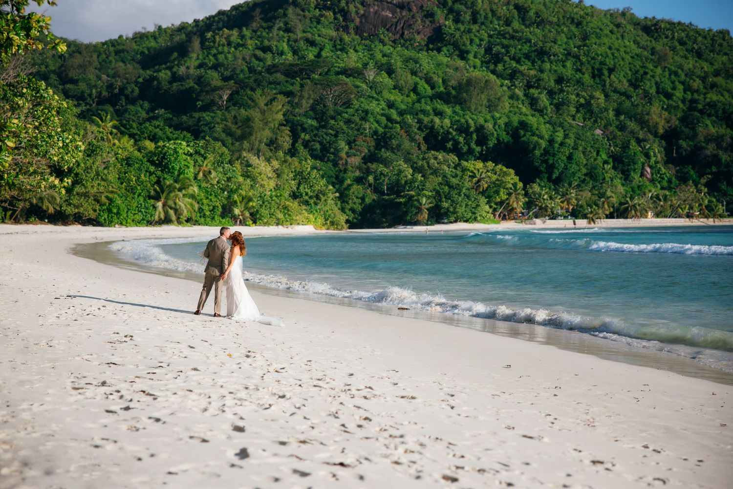 Bride and groom embracing on pristine white sand beach with turquoise water and lush hills.