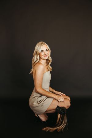 Smiling blonde woman in beige strapless dress and black boots poses seated against dark studio background.