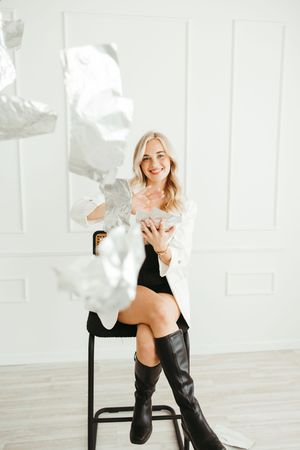 Smiling blonde woman in white blazer and black knee-high boots sits on stool with silver balloons in bright white room.