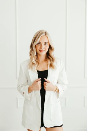 Confident blonde woman in white blazer and black outfit, professional portrait against white wall.