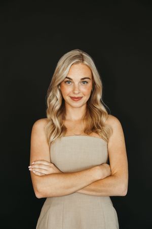 Confident blonde woman in strapless beige dress with arms crossed, smiling against dark background.