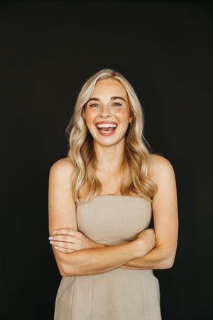 Smiling blonde woman in beige strapless dress with arms crossed against black background.