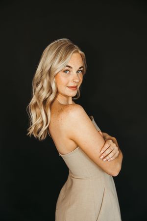 Blonde woman with freckles in beige strapless dress poses with arms crossed against black background.