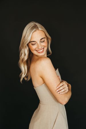 Smiling blonde woman with freckles in a beige strapless dress, eyes closed, posing against a dark background.