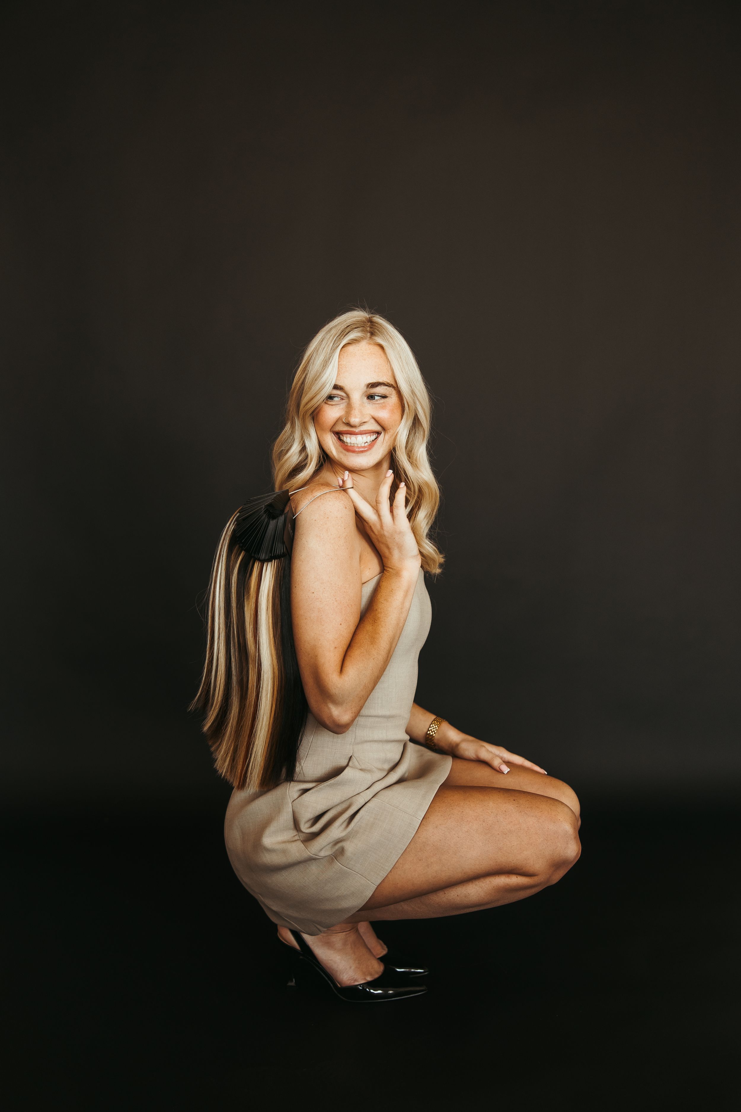 Smiling blonde woman in beige mini dress crouching against dark background, wearing gold bracelet and black heels.