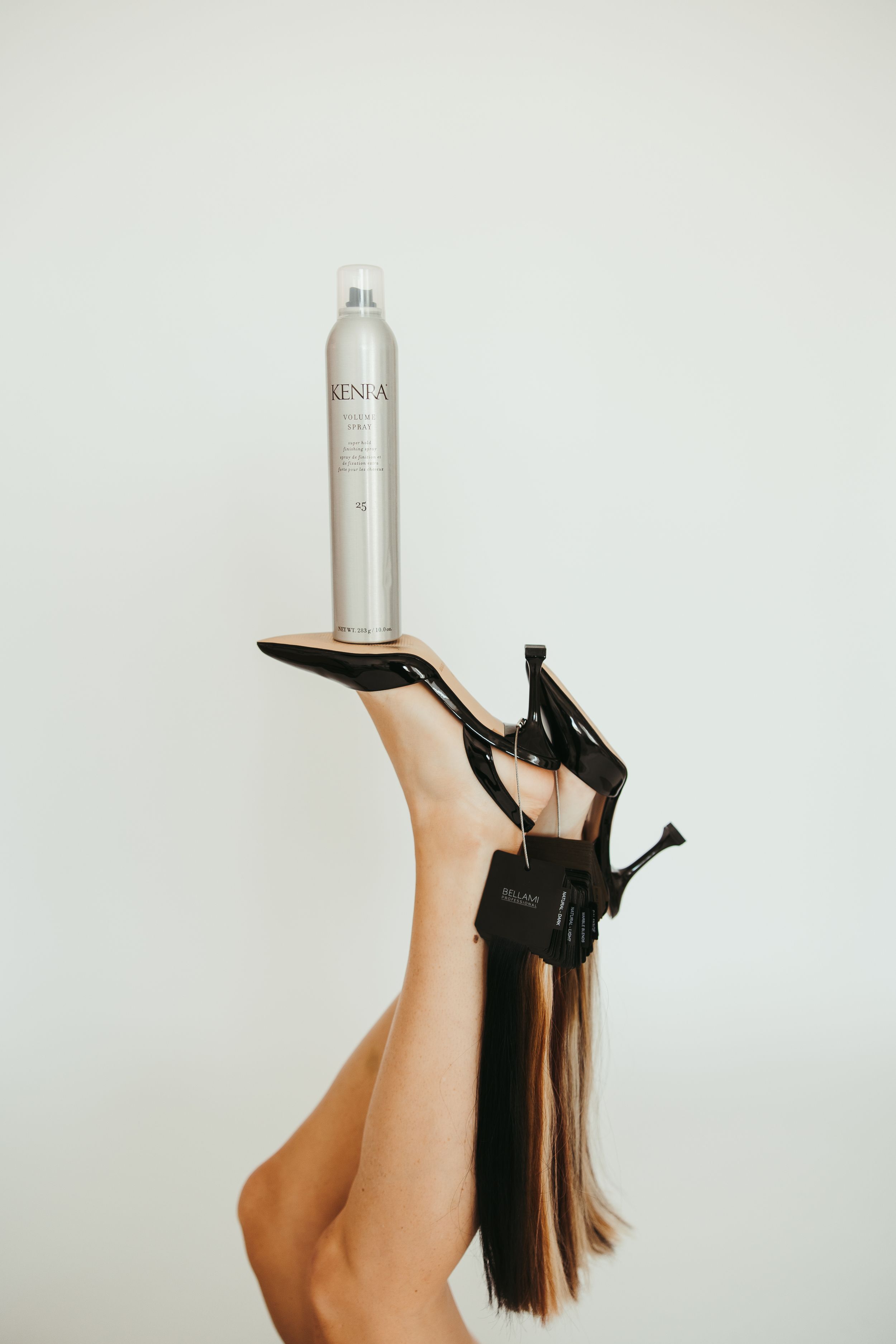 Legs raised holding black high heels with Kenra hairspray balanced on foot against white background.