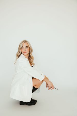 Blonde woman in white blazer and black boots crouching against a white background in a stylish pose.