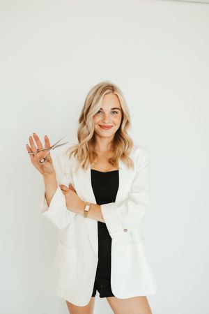 Smiling blonde woman in white blazer holding hair scissors, professional hairstylist portrait.