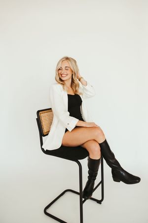 Blonde woman in white blazer and black boots sitting on chair against white background.