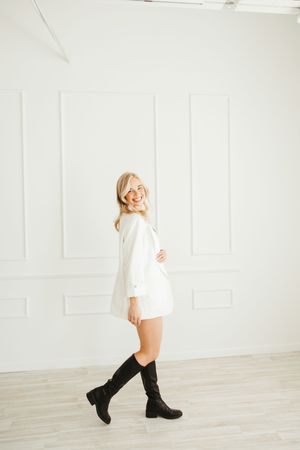 Smiling blonde woman in white mini dress and black knee-high boots posing in a bright white studio.