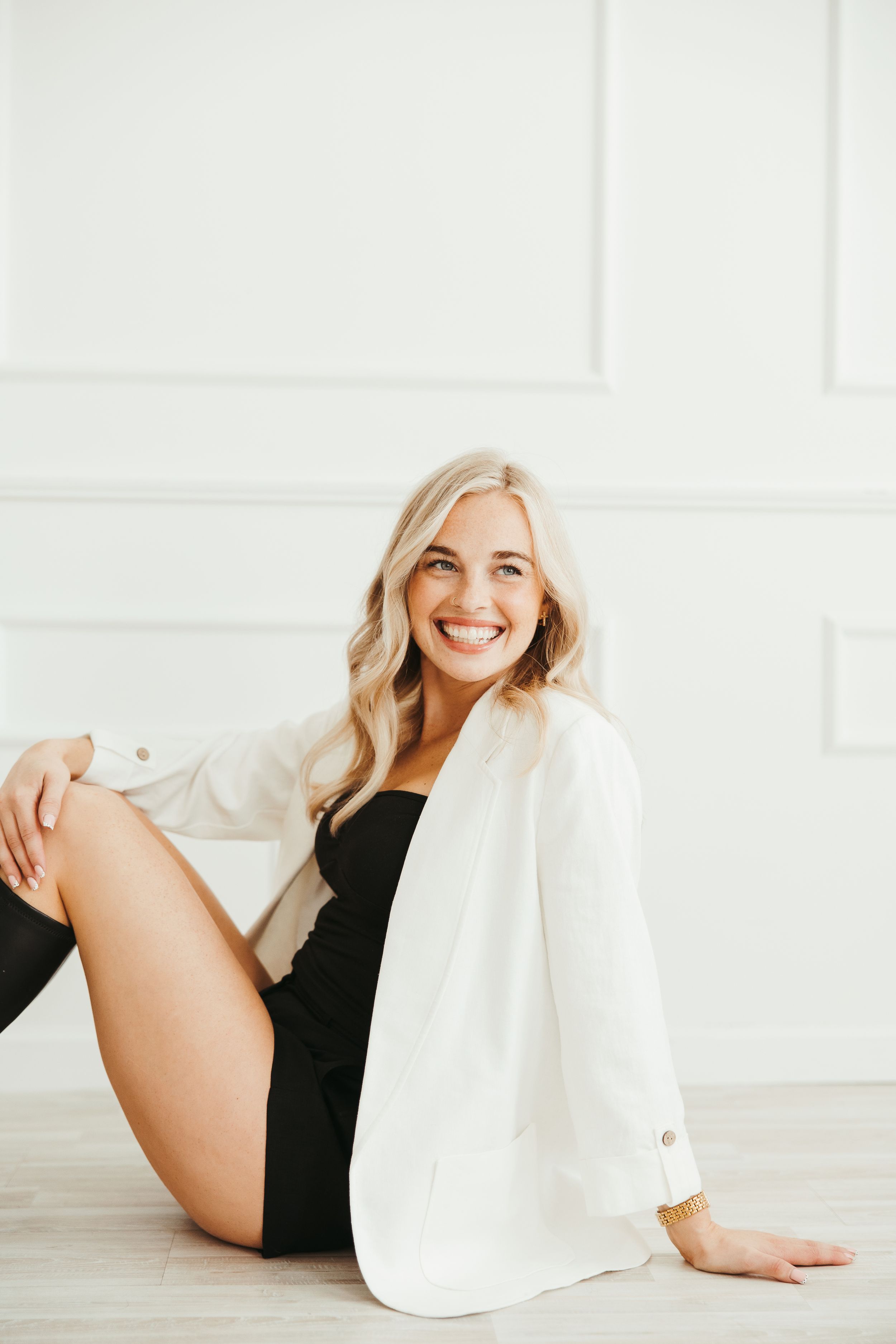Smiling blonde woman in white blazer and black outfit sitting on floor against white wall.