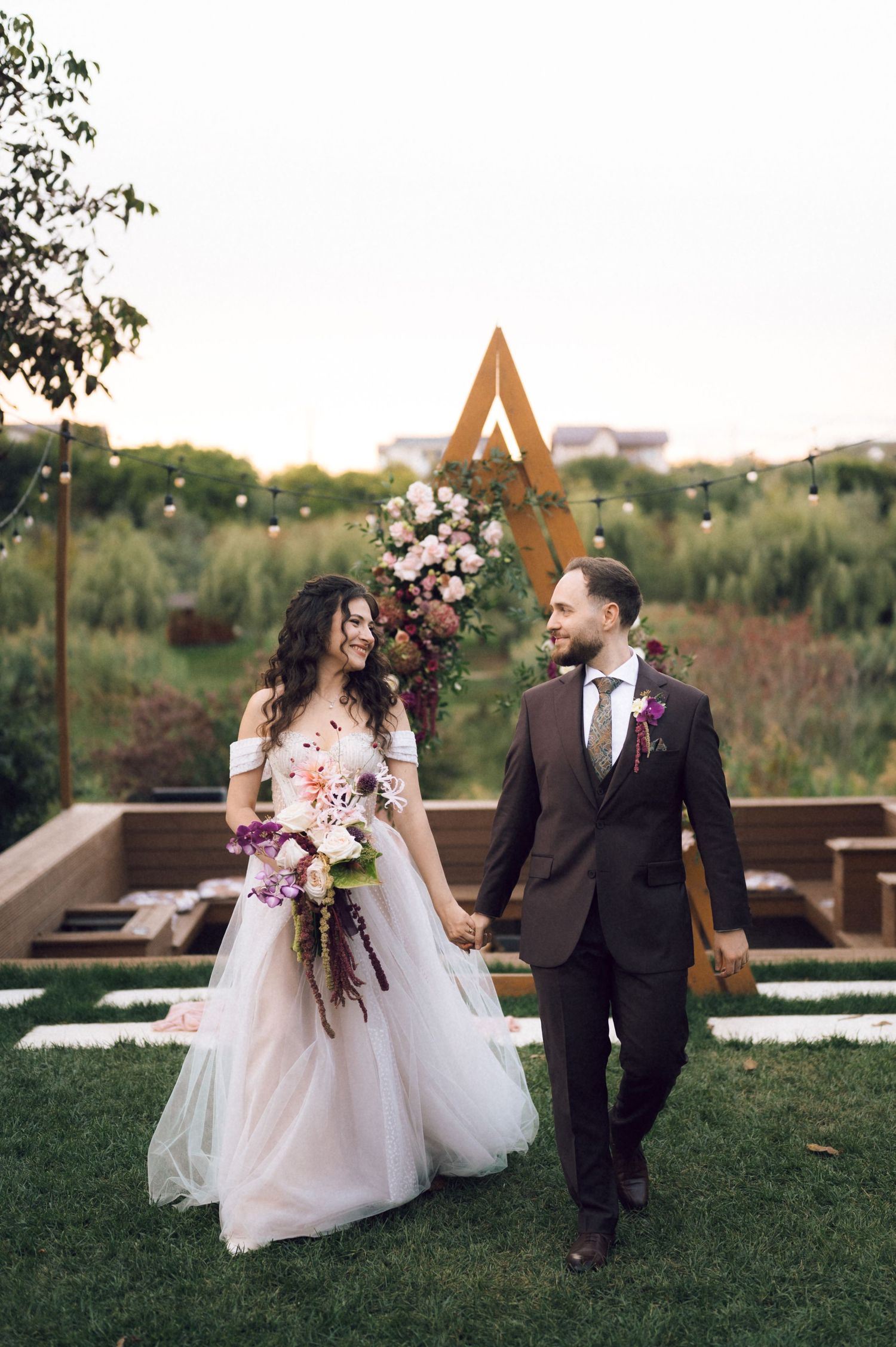 Bride and groom holding hands walking outdoors at elegant garden wedding with floral arch and string lights.