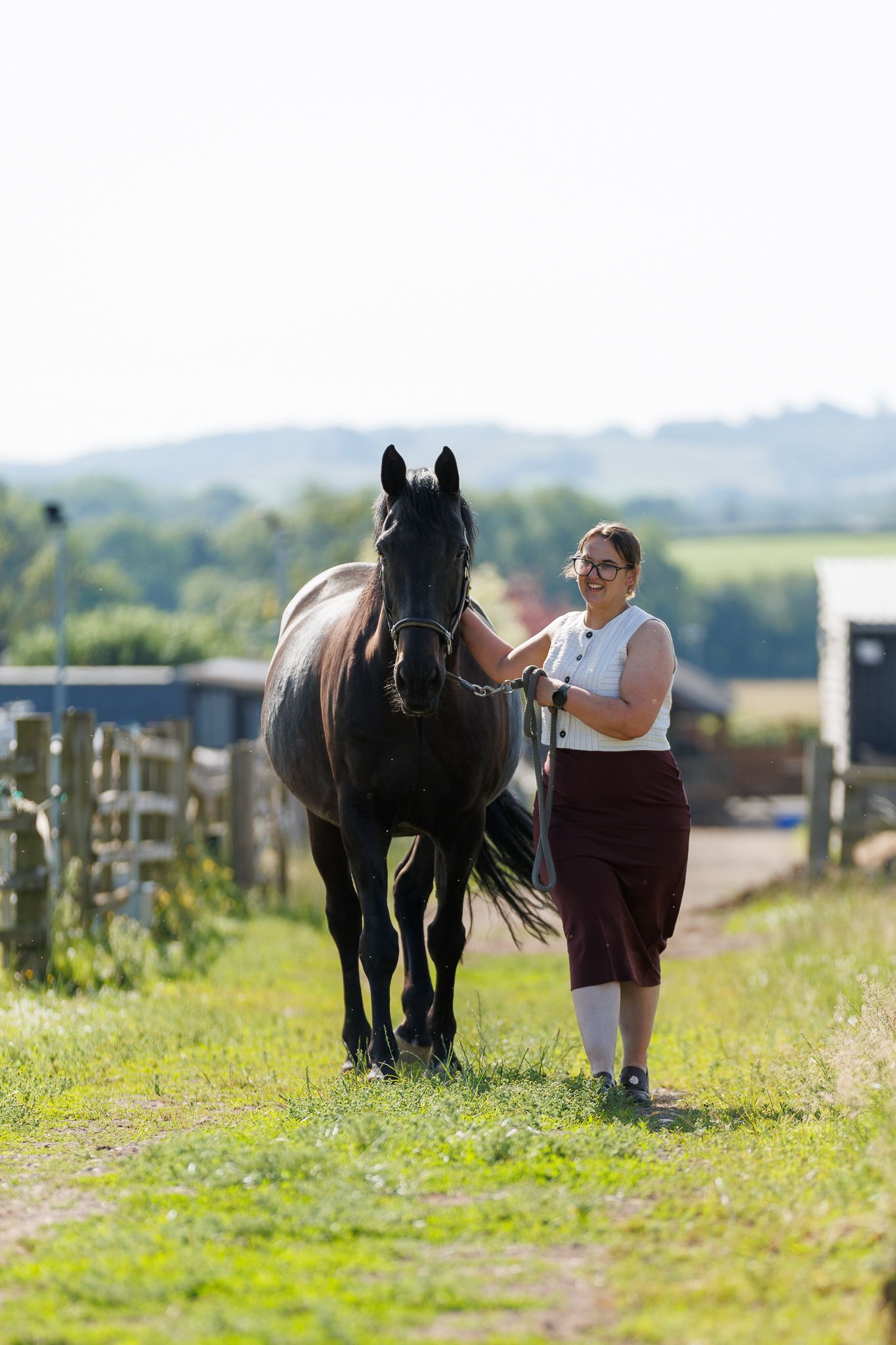 A beautiful bay horse wearing a black bridle stands next to someone in a white lace dress in an outdoor setting.