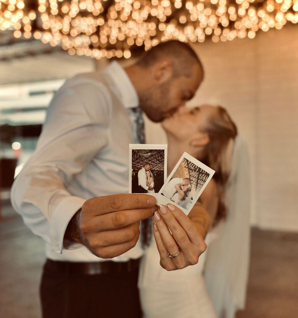Bride and groom kiss under string lights while holding two instant wedding photos in front of the camera.
