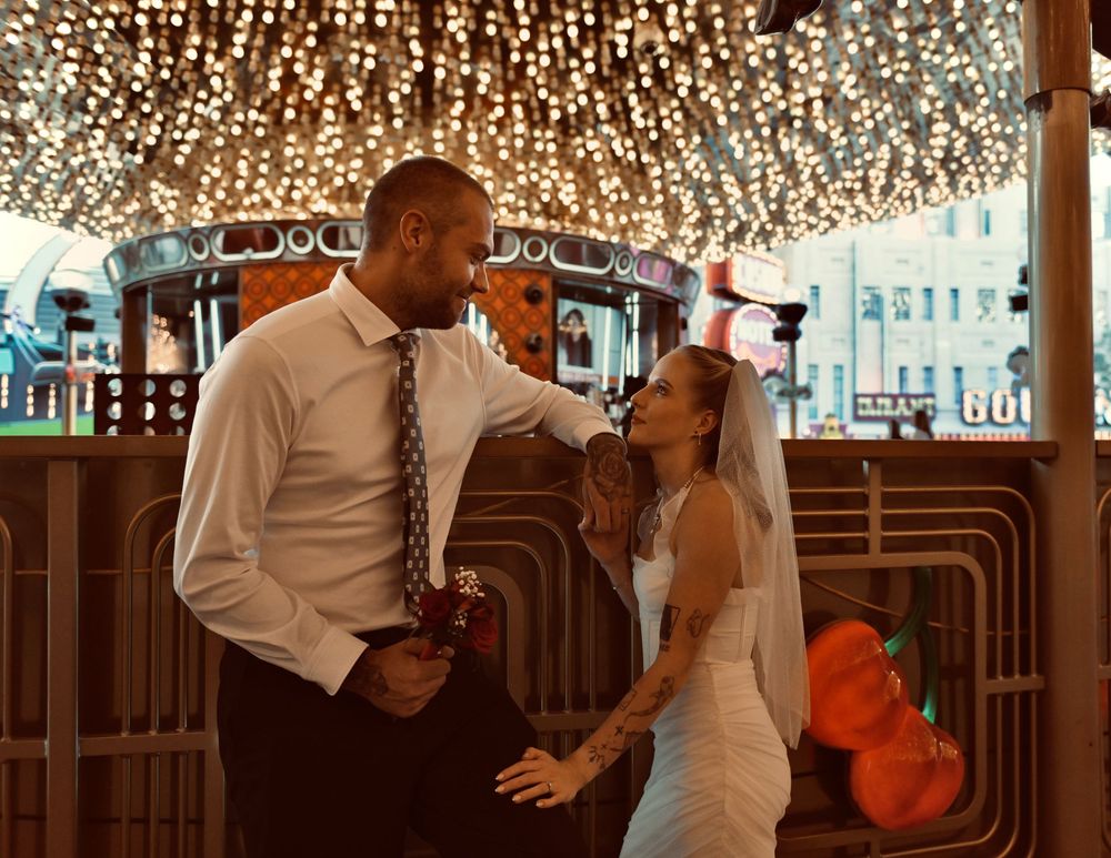 A groom in a white shirt and tie embraces a bride in a veil and white dress beneath a canopy of twinkling string lights.