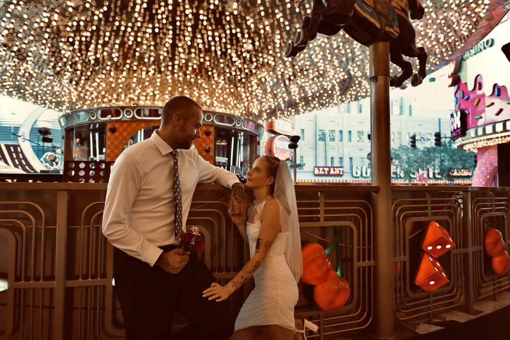 Bride in a white dress and veil embraces a groom in a white shirt and tie beneath twinkling lights on a colorful Las Vegas-style street.