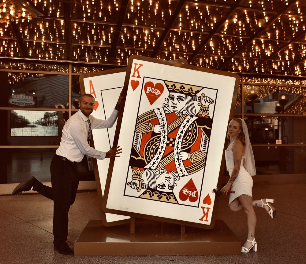 Groom and bride in a white dress pose beside a giant King of Hearts playing card under twinkling lights at a Las Vegas wedding.